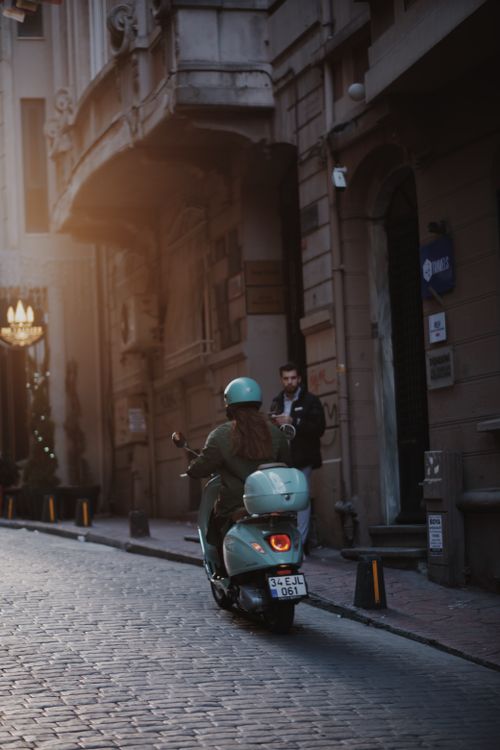 Free A woman rides a scooter through a historic city street at dusk, blending modern mobility with vintage charm. Stock Photo