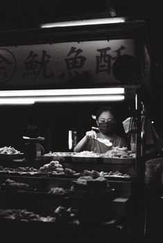 Street food vendor at night under illuminated stall, captured in monochrome, selling Asian cuisine.