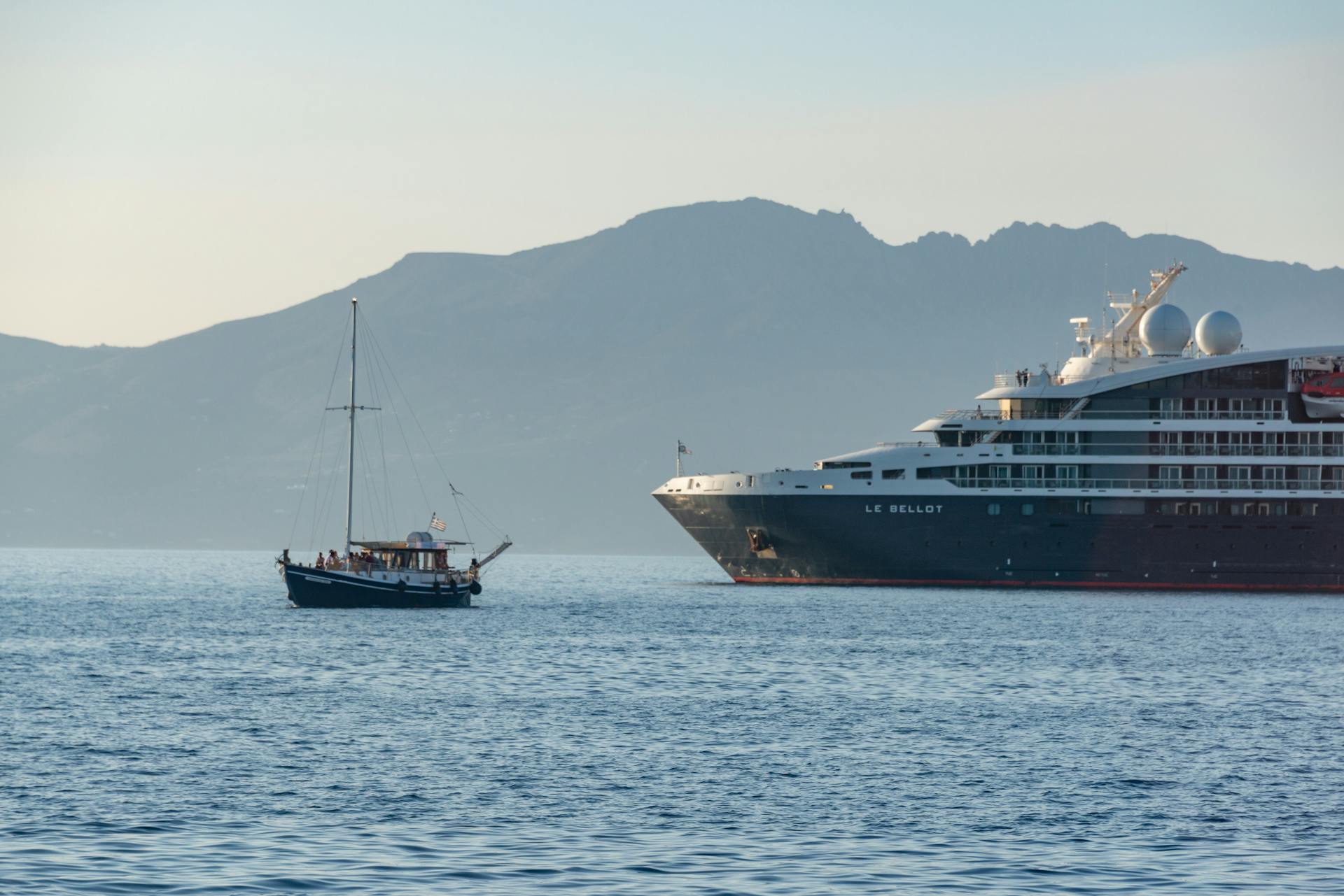 A luxurious cruise ship and a sailboat on a serene sea with mountainous background.