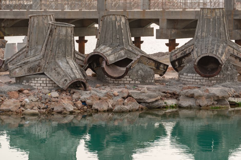 Concrete bridge construction site with rusty components and water reflection.