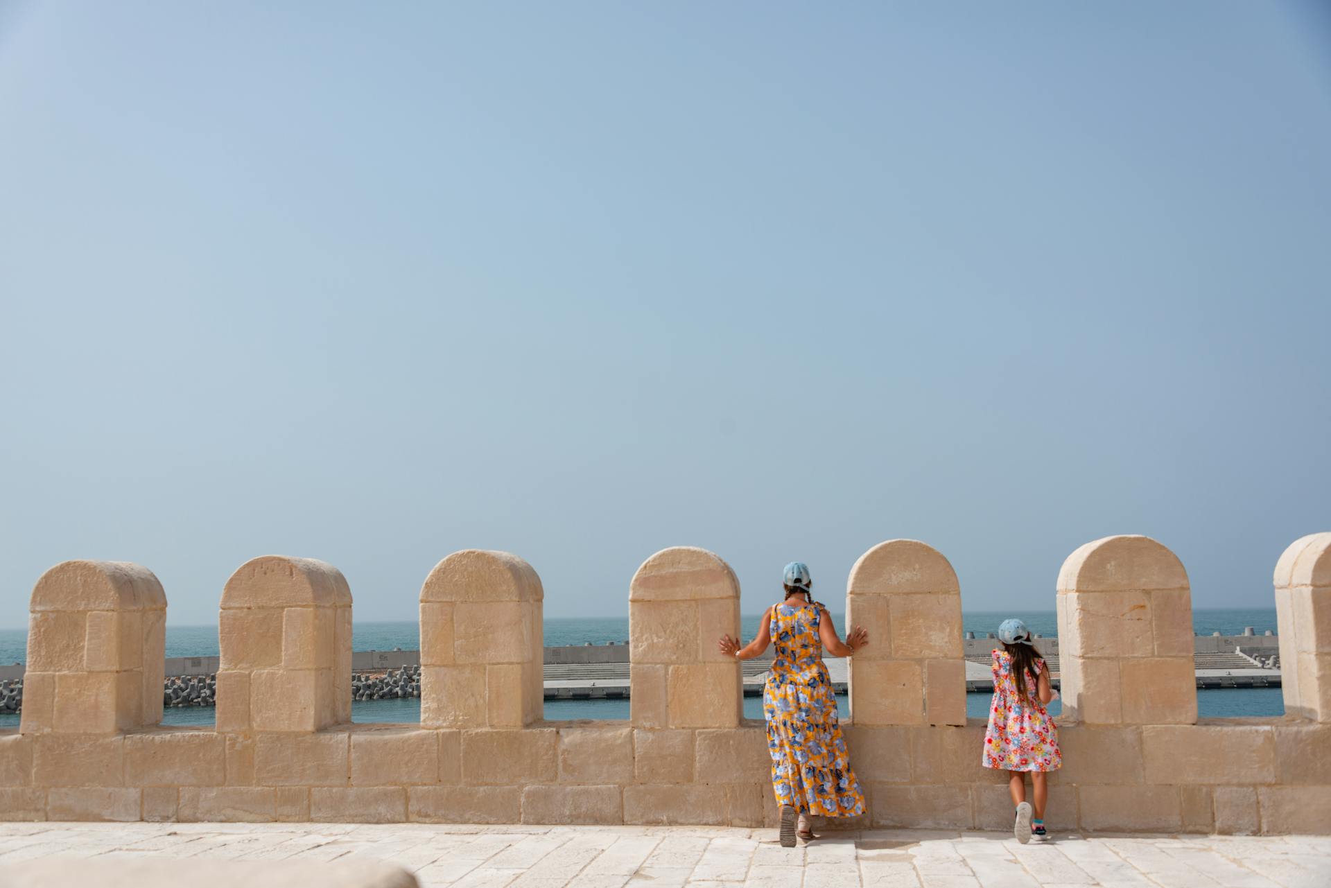 A woman and child view the sea from historic citadel walls under a clear sky.