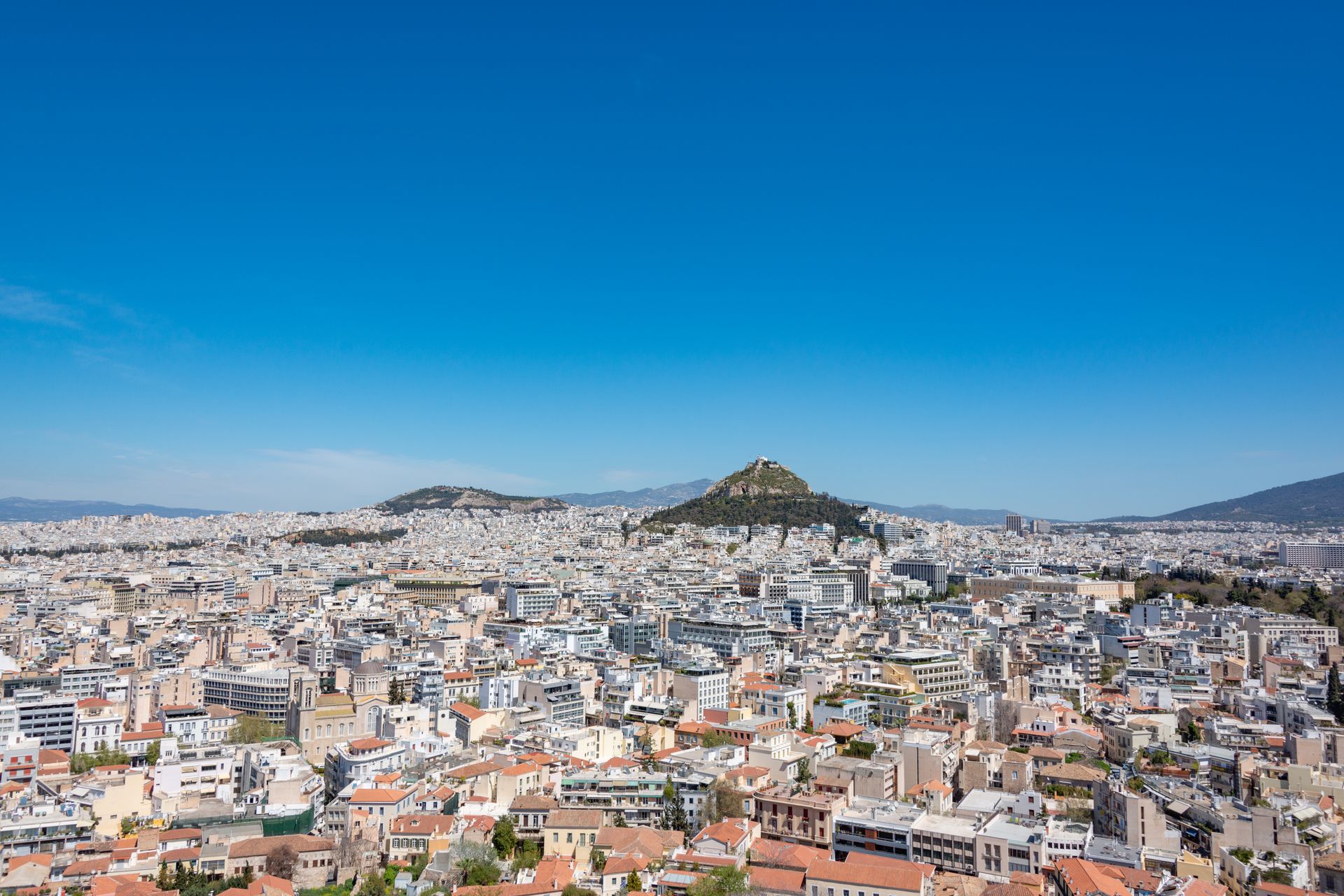A panoramic cityscape view of Athens featuring Lycabettus Hill under a clear blue sky.