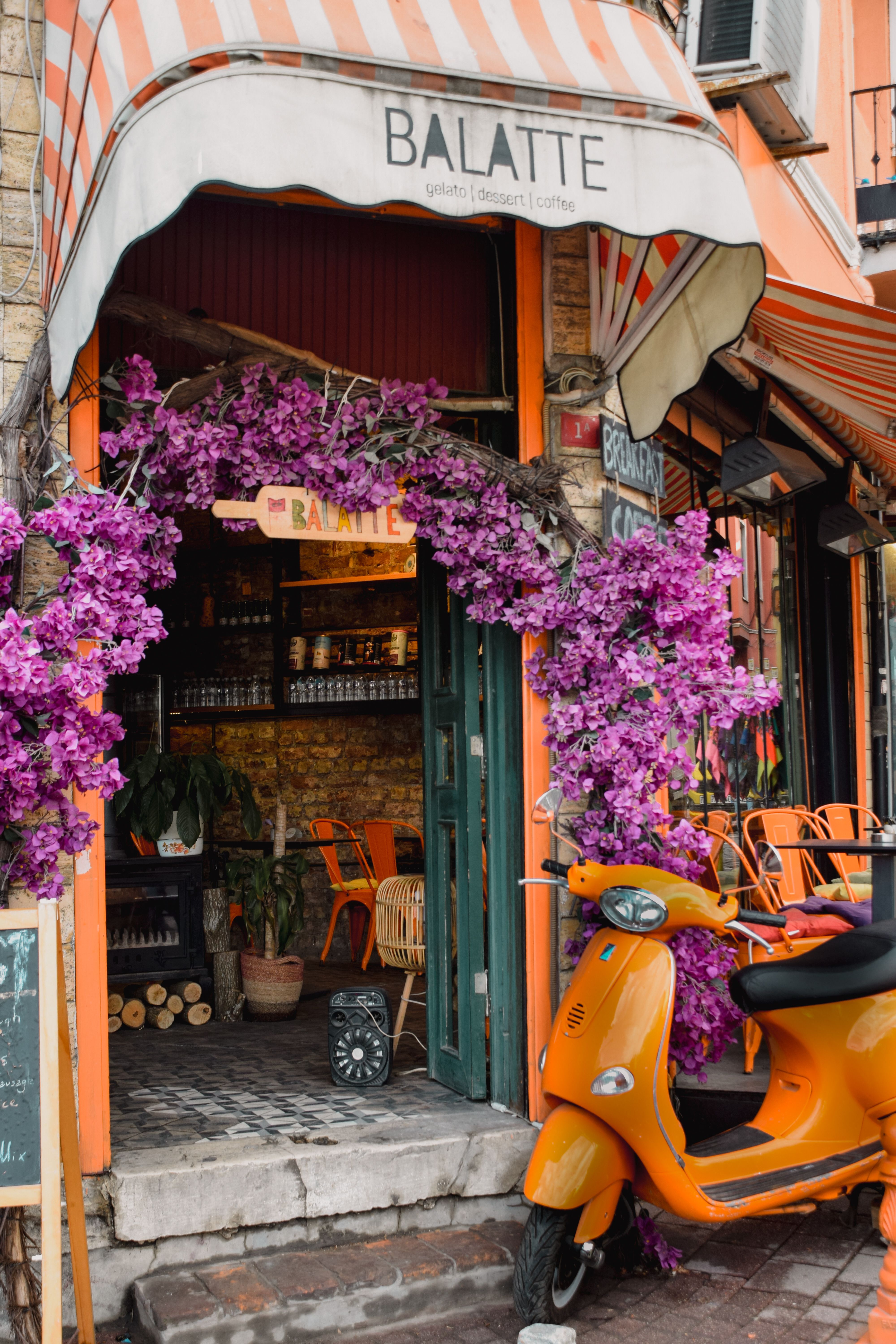 Urban café entrance with orange scooter and vibrant purple flowers under striped sunshade.