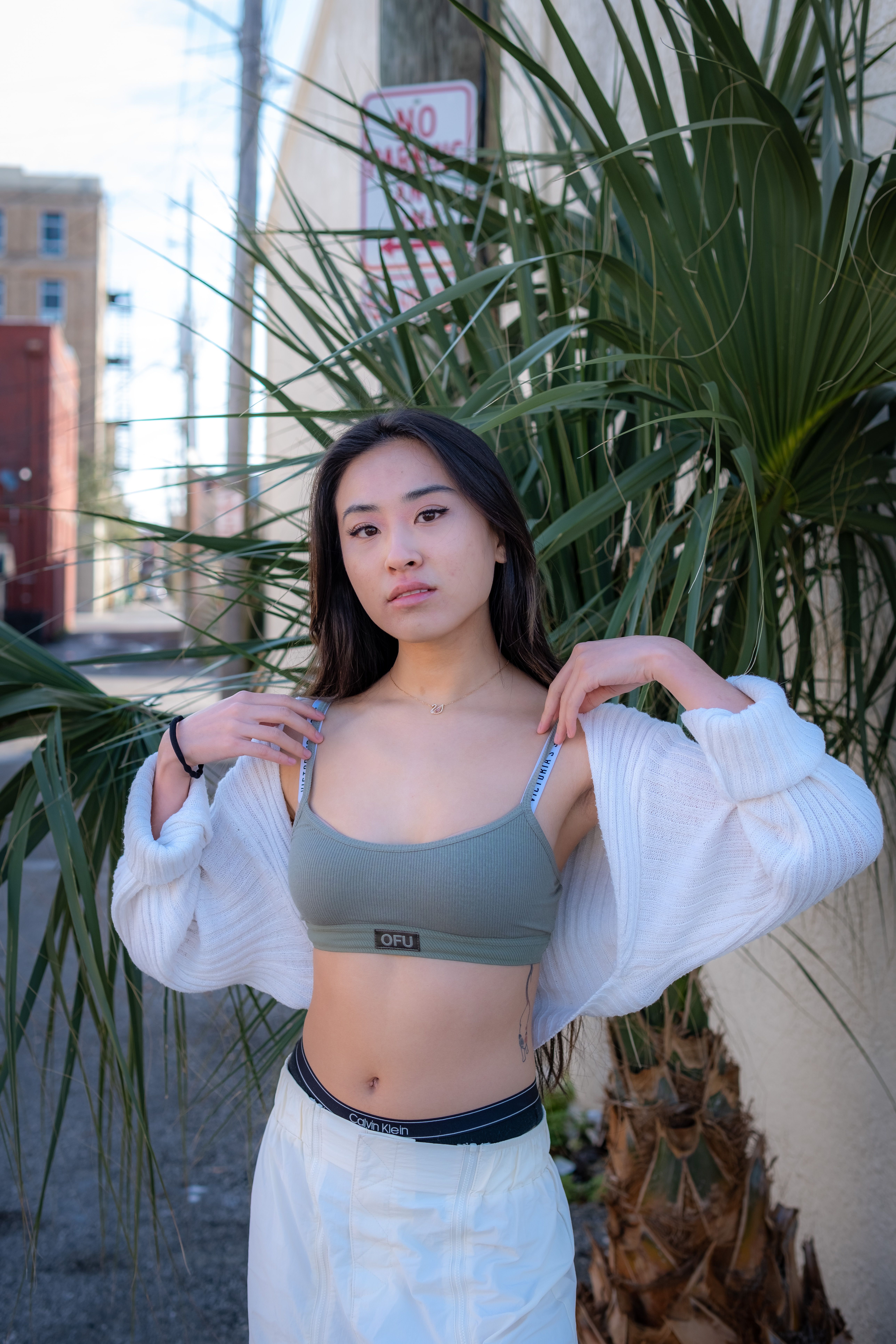 Woman in sportswear and cardigan posing outdoors by palm tree.