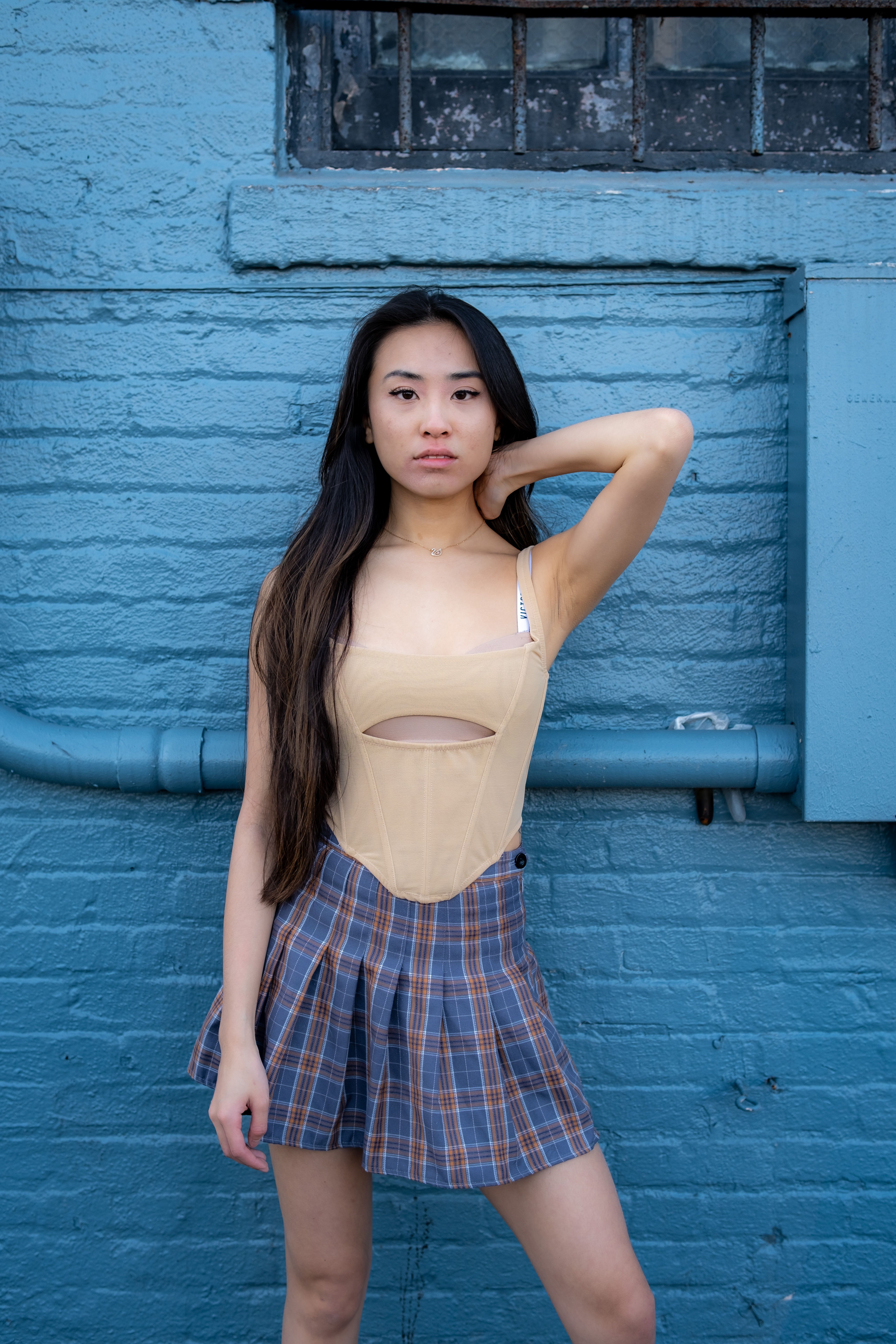 Stylish young woman in a plaid skirt and beige top posing confidently against a blue wall.