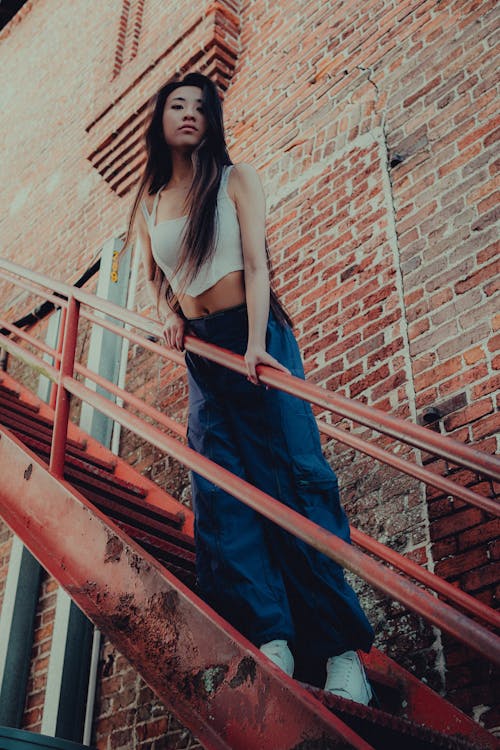 Woman in White Crop Top and Blue Pants Standing on Stairs in Low Angle Shot Photography 