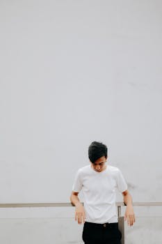 A young man in a white t-shirt stands against a minimalist urban backdrop.