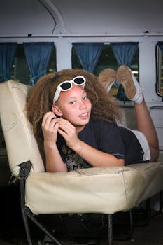 Teen girl with curly hair, sunglasses in hand, lying in a vintage bus interior, creating a relaxed travel vibe.