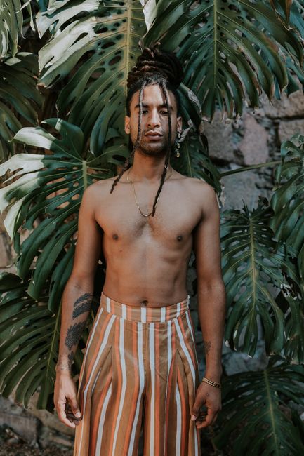 Young Hispanic man with tattoos and dreadlocks standing by tropical leaves, exuding natural style.