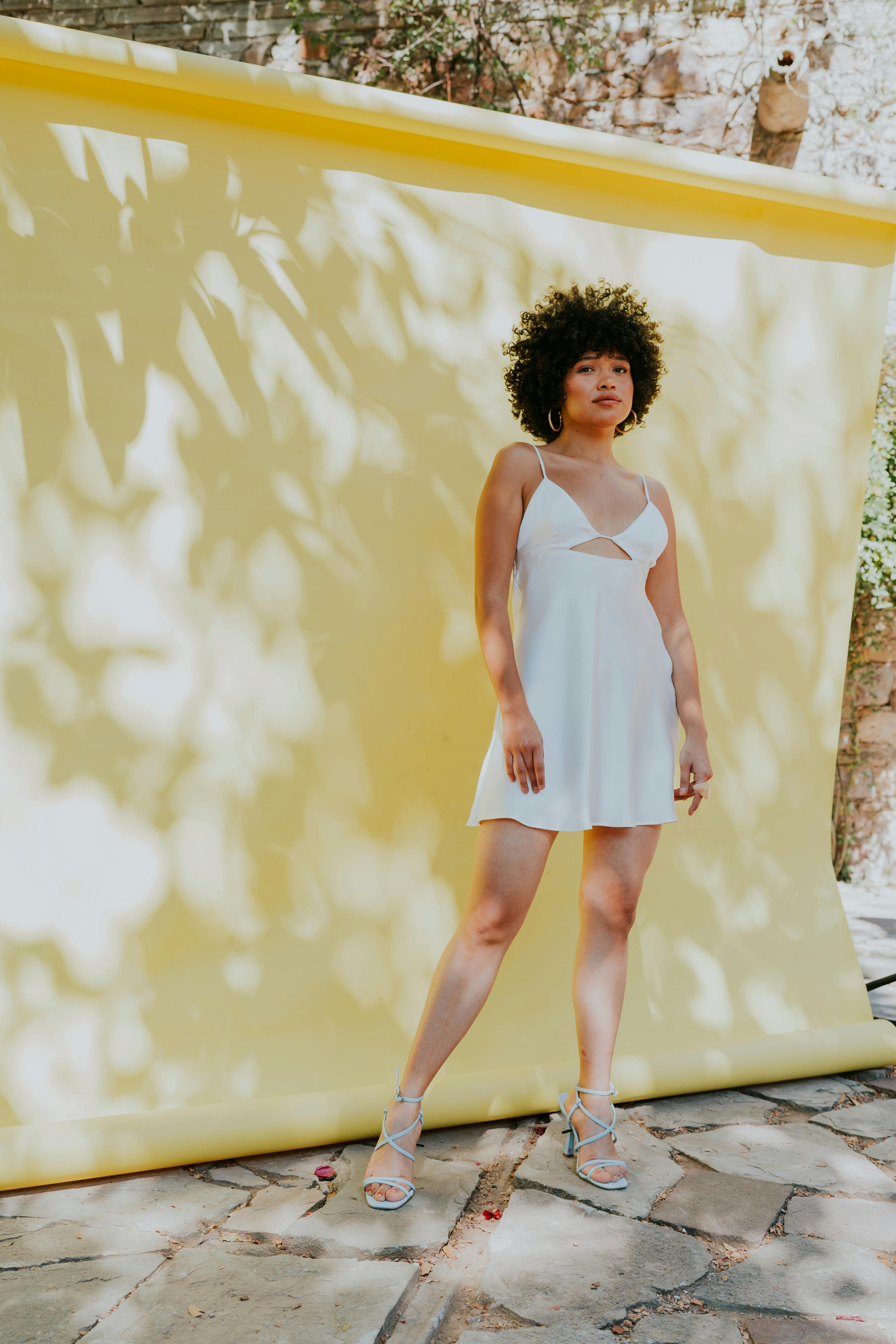 Young woman in white dress posing against yellow backdrop in sunlight.