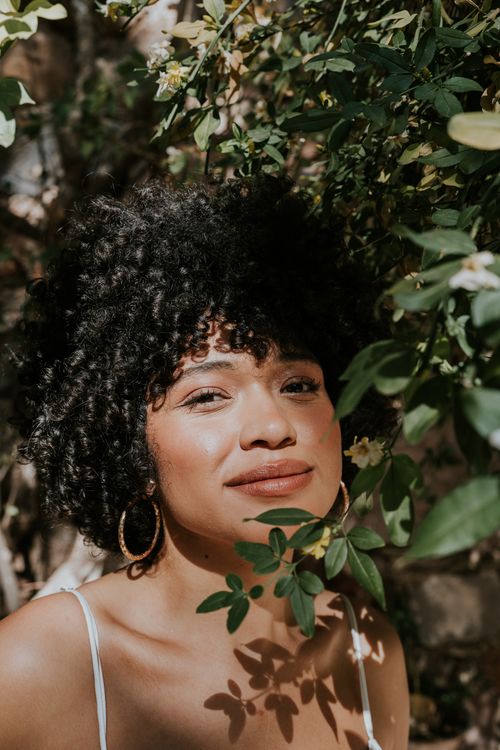 Free A joyful woman with curly hair posing amidst lush greenery on a sunny day outdoors. Stock Photo