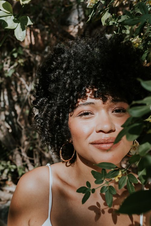 Free Beautiful woman with curly hair smiling amidst lush green leaves in sunlight. Stock Photo