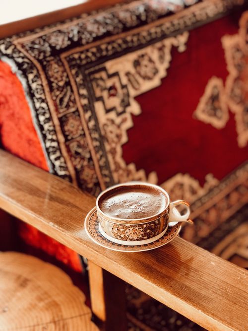 Free A beautifully patterned coffee cup sits on a wooden armrest against a rich, ornate rug backdrop. Stock Photo