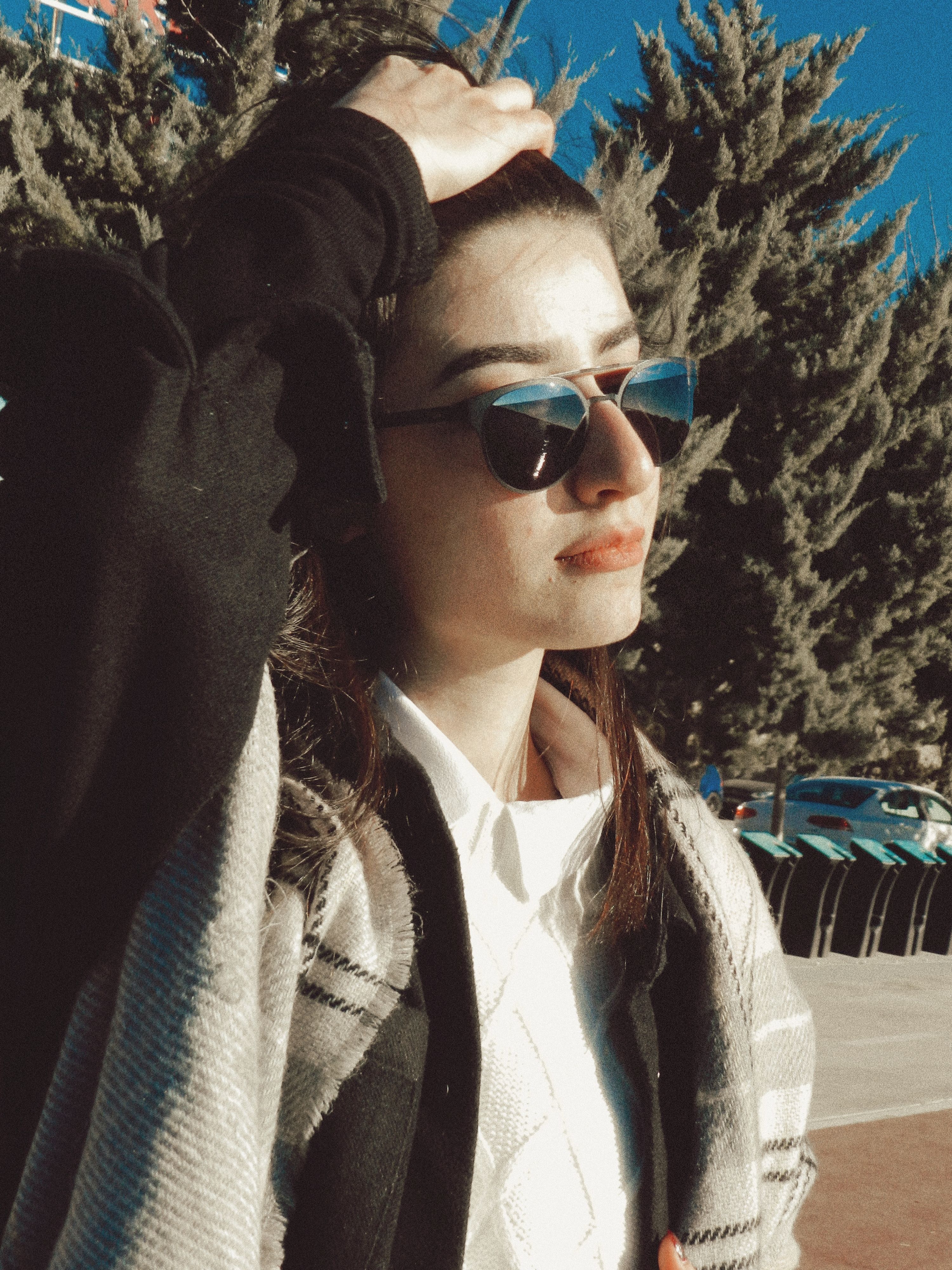 A fashionable woman wearing sunglasses stands outdoors, basking in the sun with hand on head.