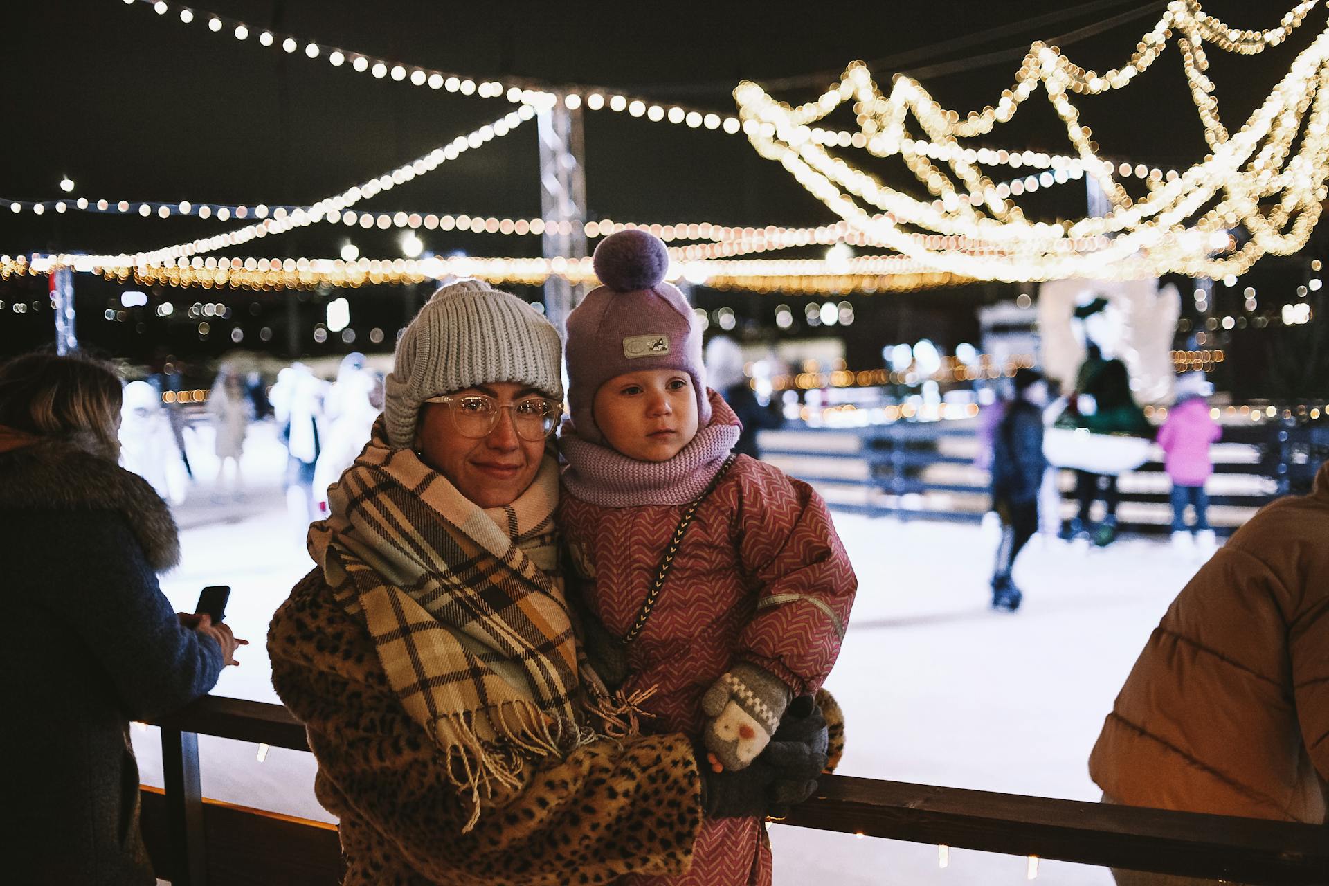 A mother and child enjoying a winter evening at a festive outdoor ice rink with decorative lights.