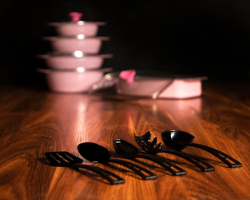 Close-up of Black Cooking Utensils Lying on a Wooden Table with Pink Pots in the Background 