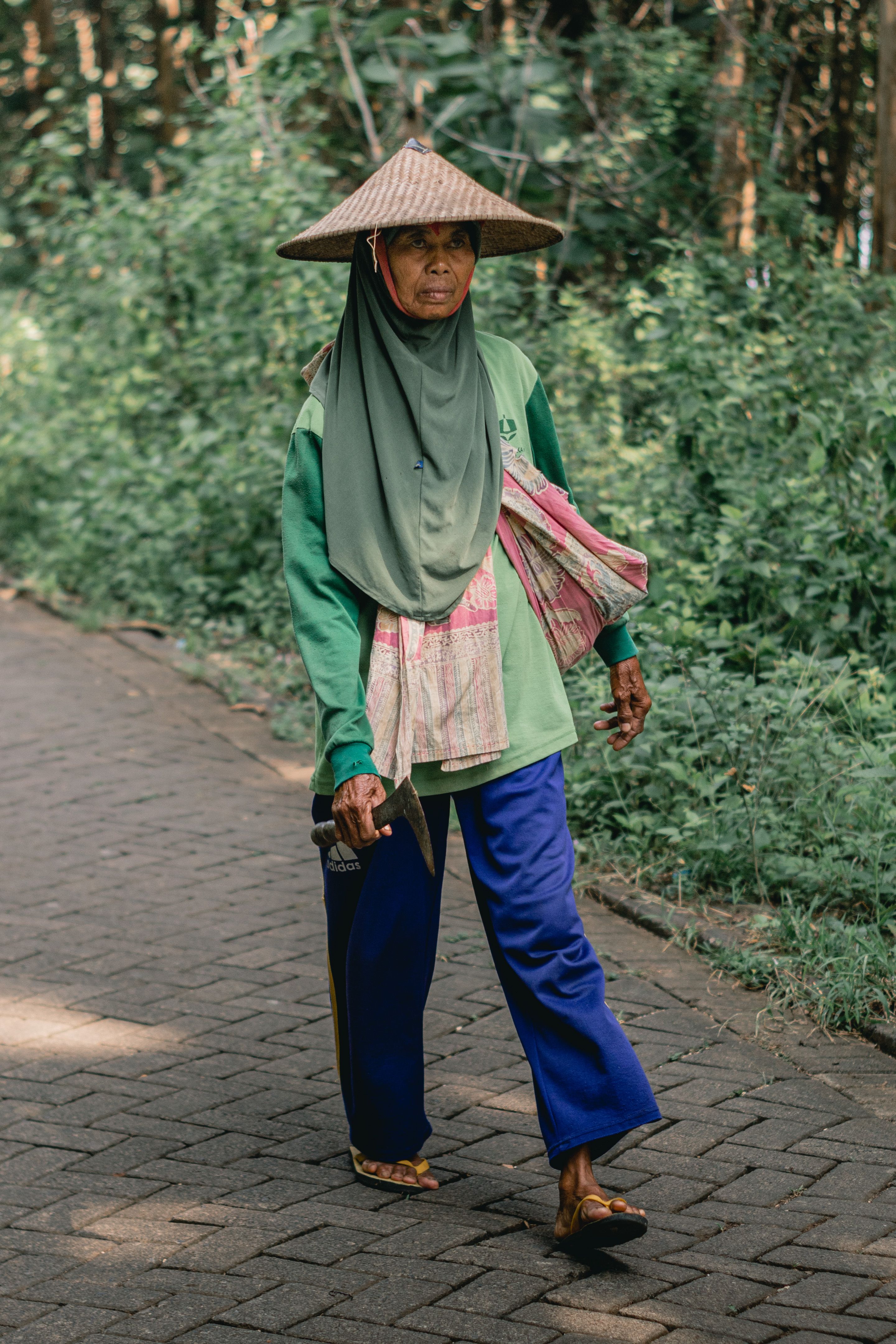 An elderly woman walks on a forest path in Brondong, Indonesia, wearing traditional attire.