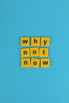 Yellow letter tiles spell 'why not now' on a vibrant blue backdrop, creating a motivational scene.