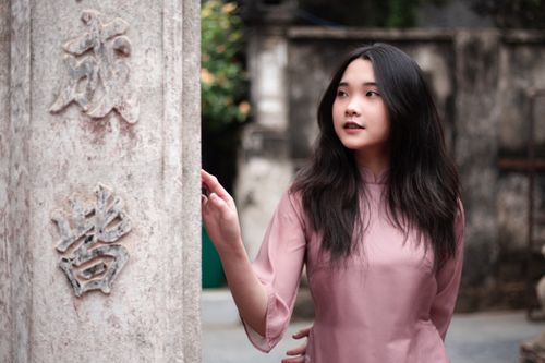 Free A young Asian woman in traditional attire poses beside an intricately carved stone wall outdoors. Stock Photo
