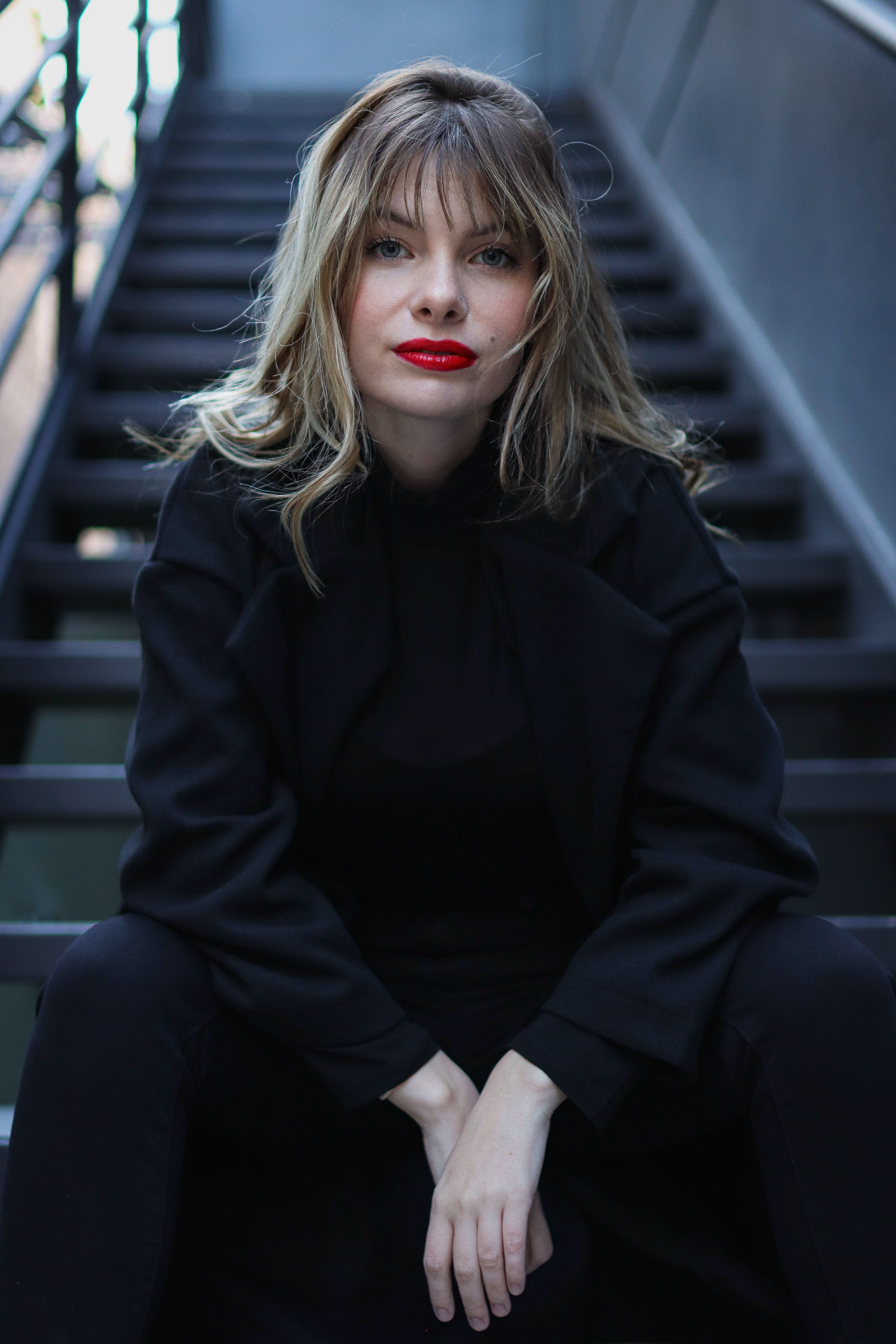Fashionable woman in dark clothing sitting on outdoor staircase with red lipstick.