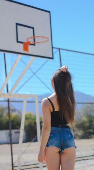A woman stands on an outdoor basketball court during a sunny day, looking at the hoop.