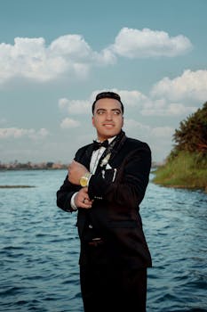 Portrait of a confident man in a black suit posing by a peaceful water setting.
