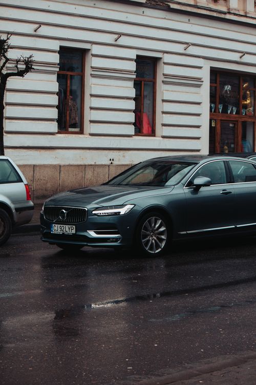 Free A Volvo sedan parked on a wet city street in front of a historic building, creating a modern meets classic vibe. Stock Photo