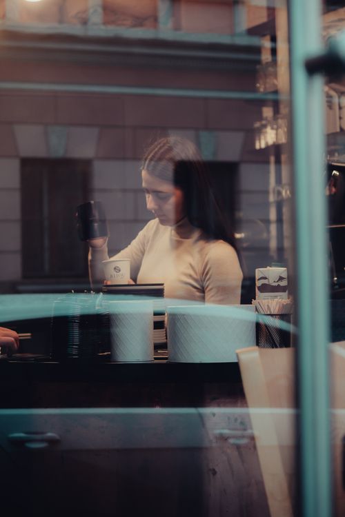 Free A woman making coffee at a café counter viewed through a window, creating a cozy urban setting. Stock Photo