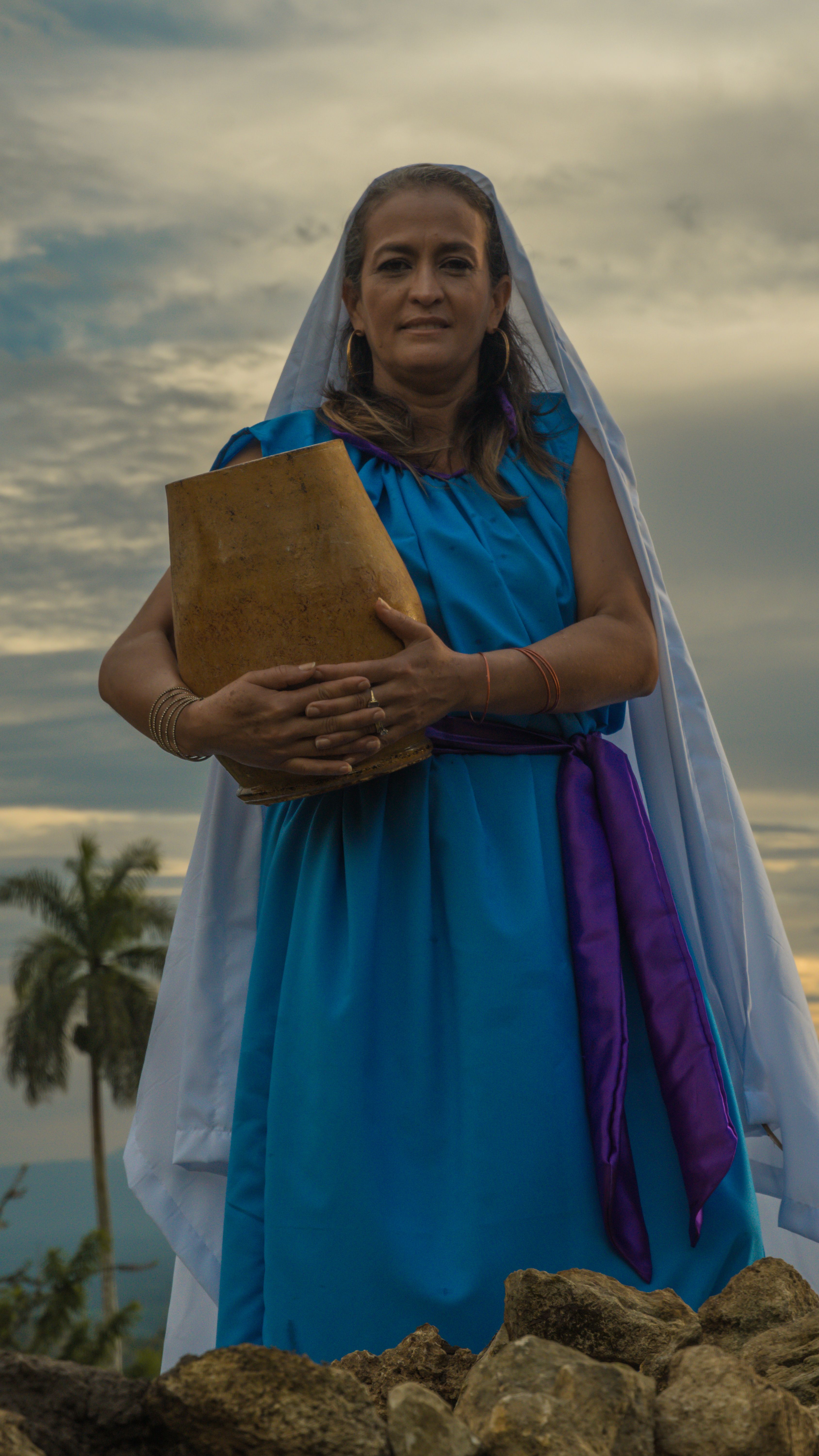 A woman in traditional clothing holds a wooden tablet against a scenic backdrop outdoors.