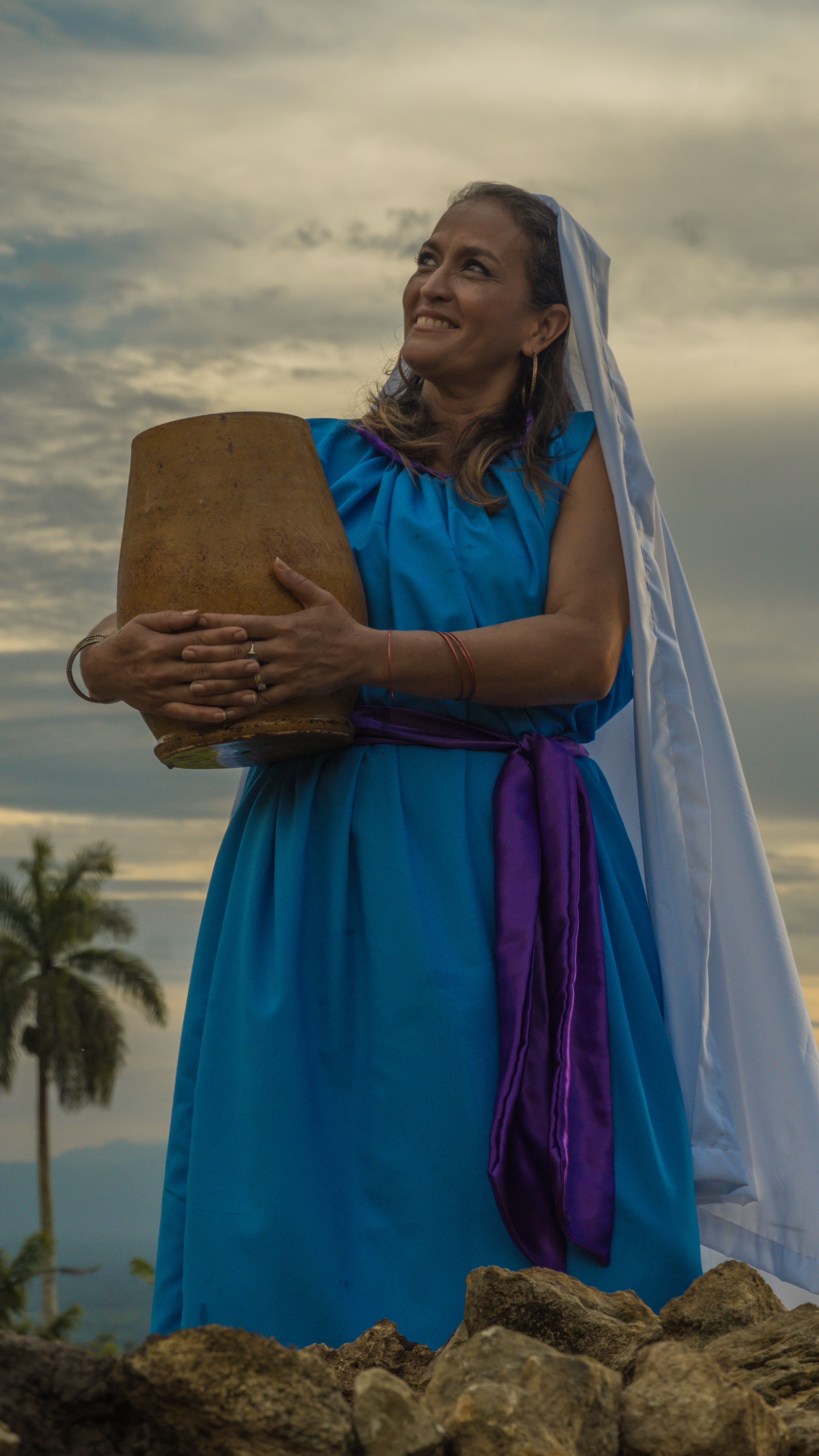 Smiling woman in blue dress with a veil holding clay pot outdoors against a cloudy sky.