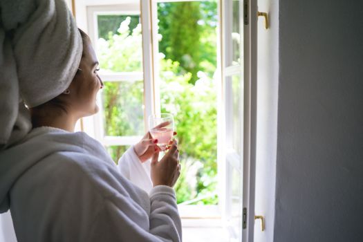 Woman in bathrobe enjoying a drink by the window, embracing a tranquil morning moment.