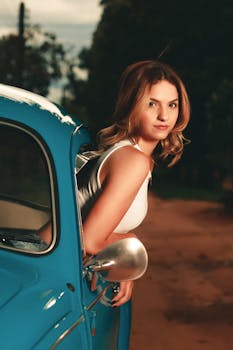 A young woman in a tank top leans out of a blue vintage car, captured in an outdoor setting.
