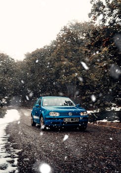 A blue car navigates a snowy road through a forest, capturing winter adventure.