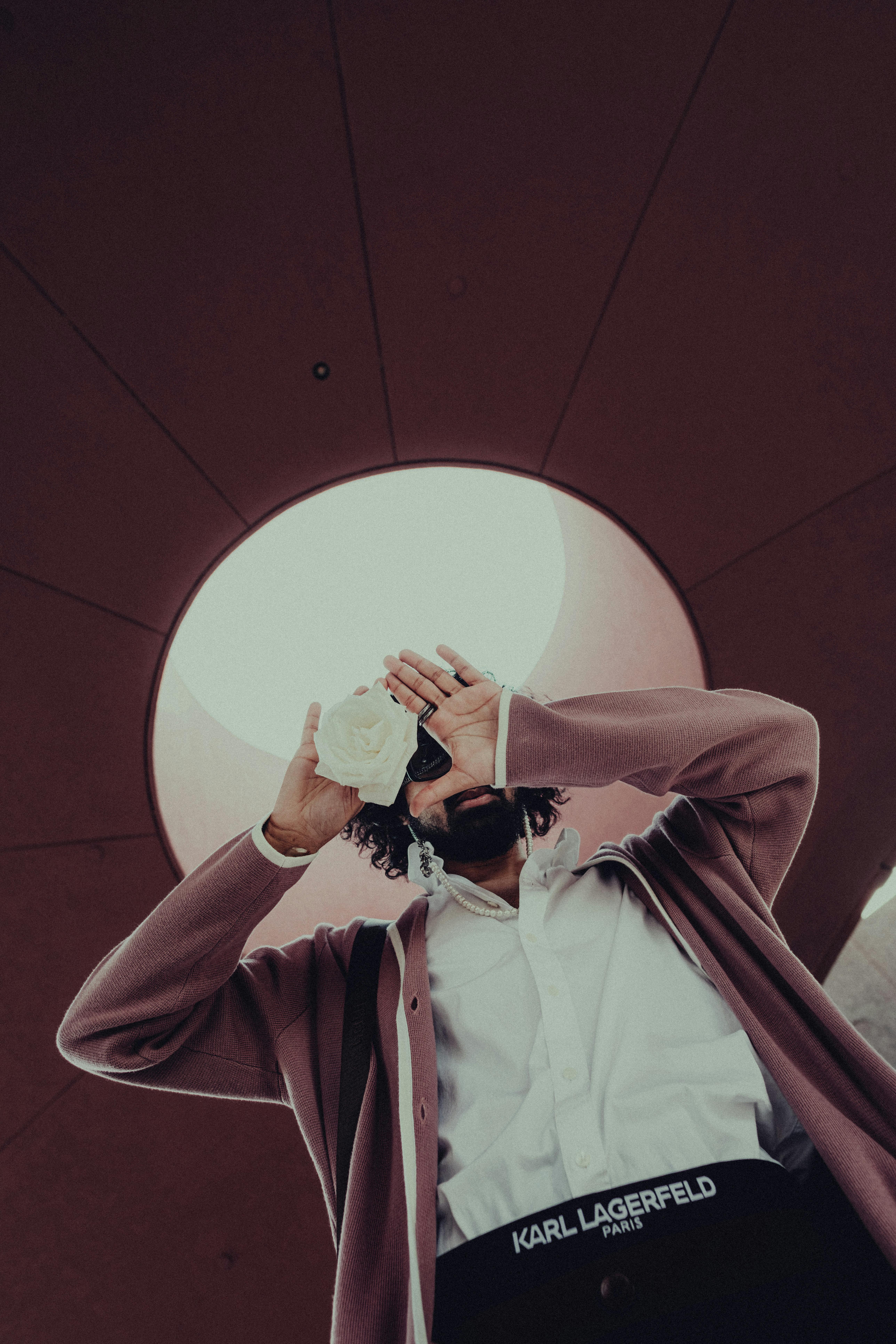 A stylish man in a pink blazer poses under a skylight, holding a white rose.