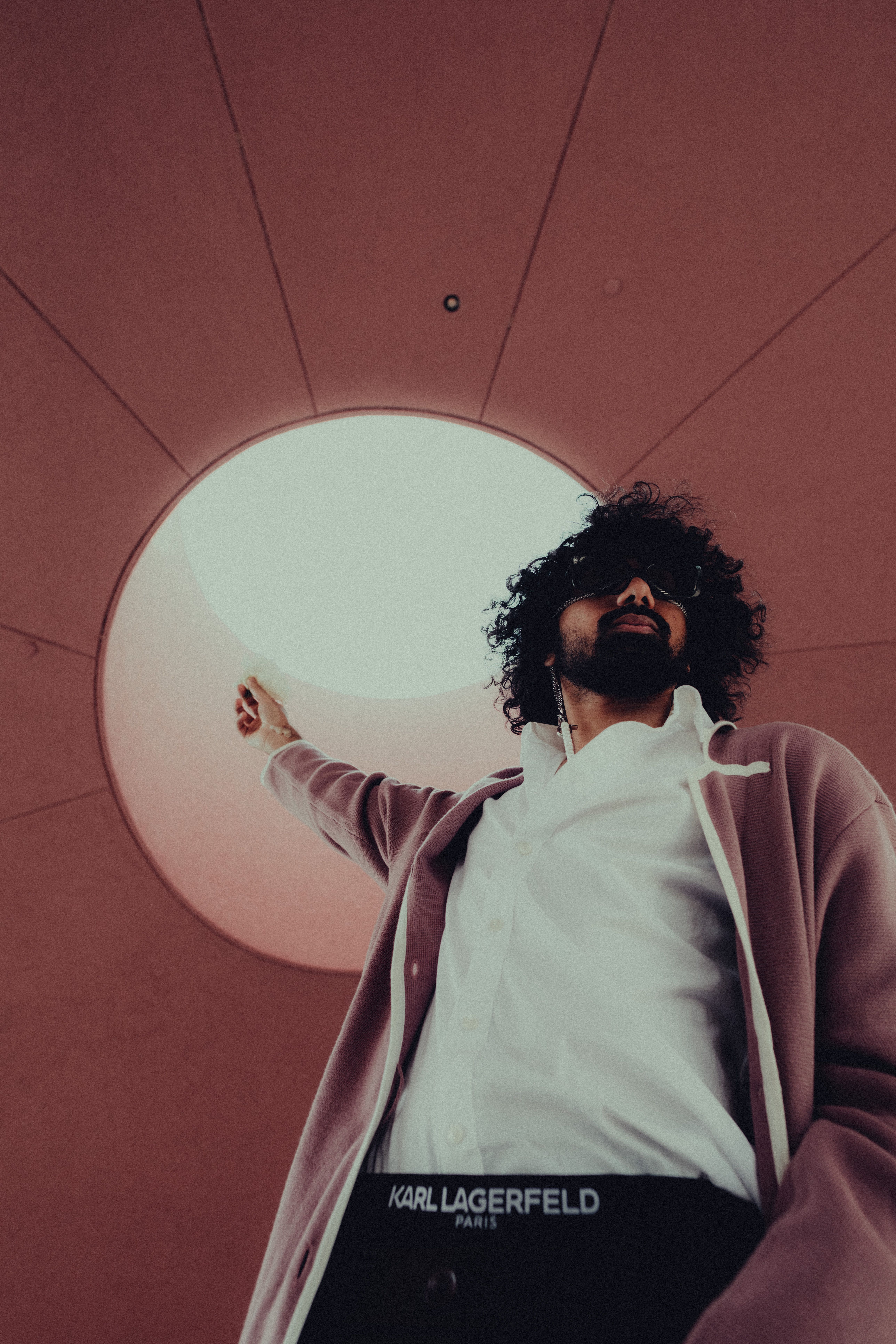Man with curly hair and sunglasses poses stylishly under a round skylight, wearing a fashionable pink jacket.