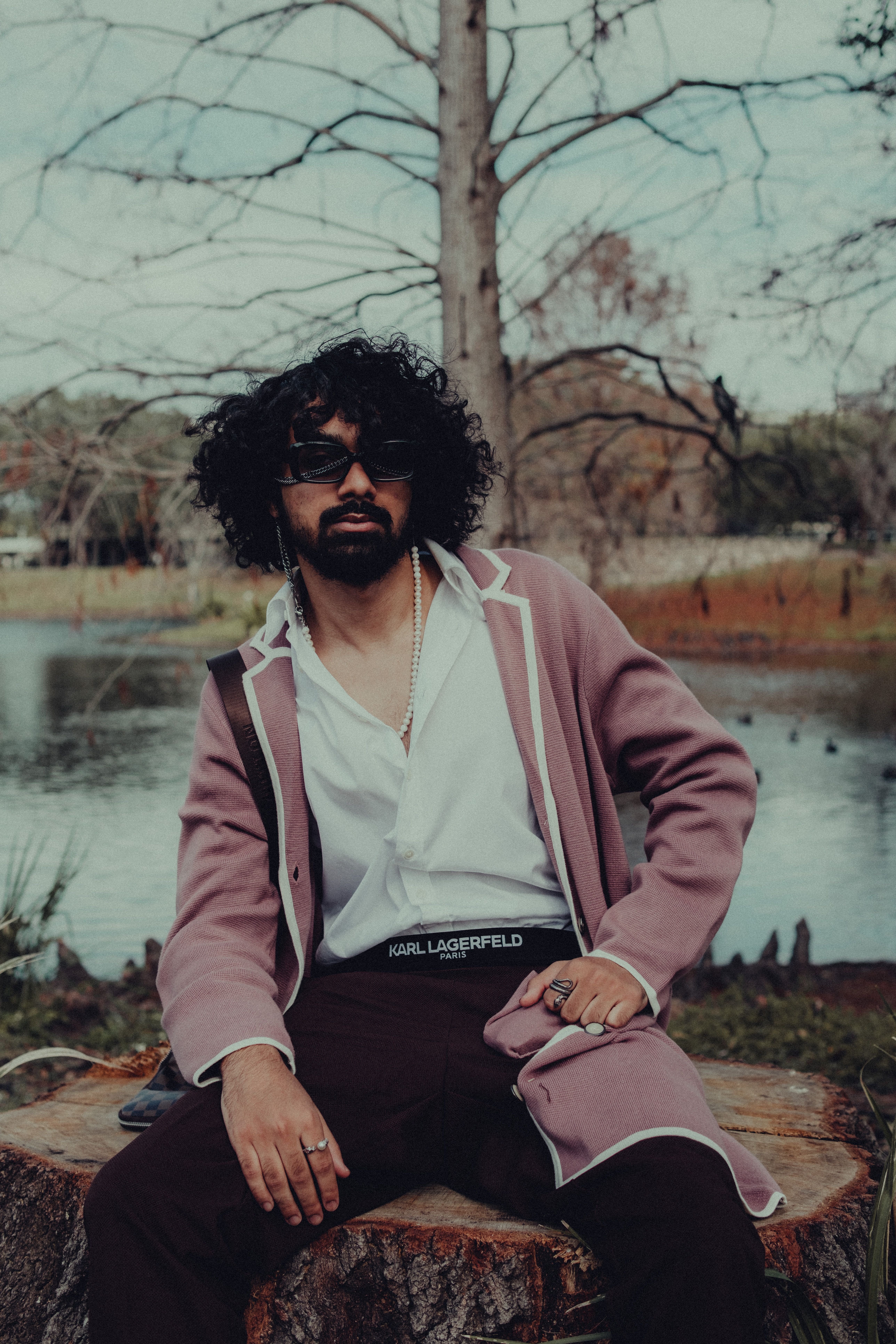 Fashionable man with sunglasses posing by a lakeside in a fall park setting.