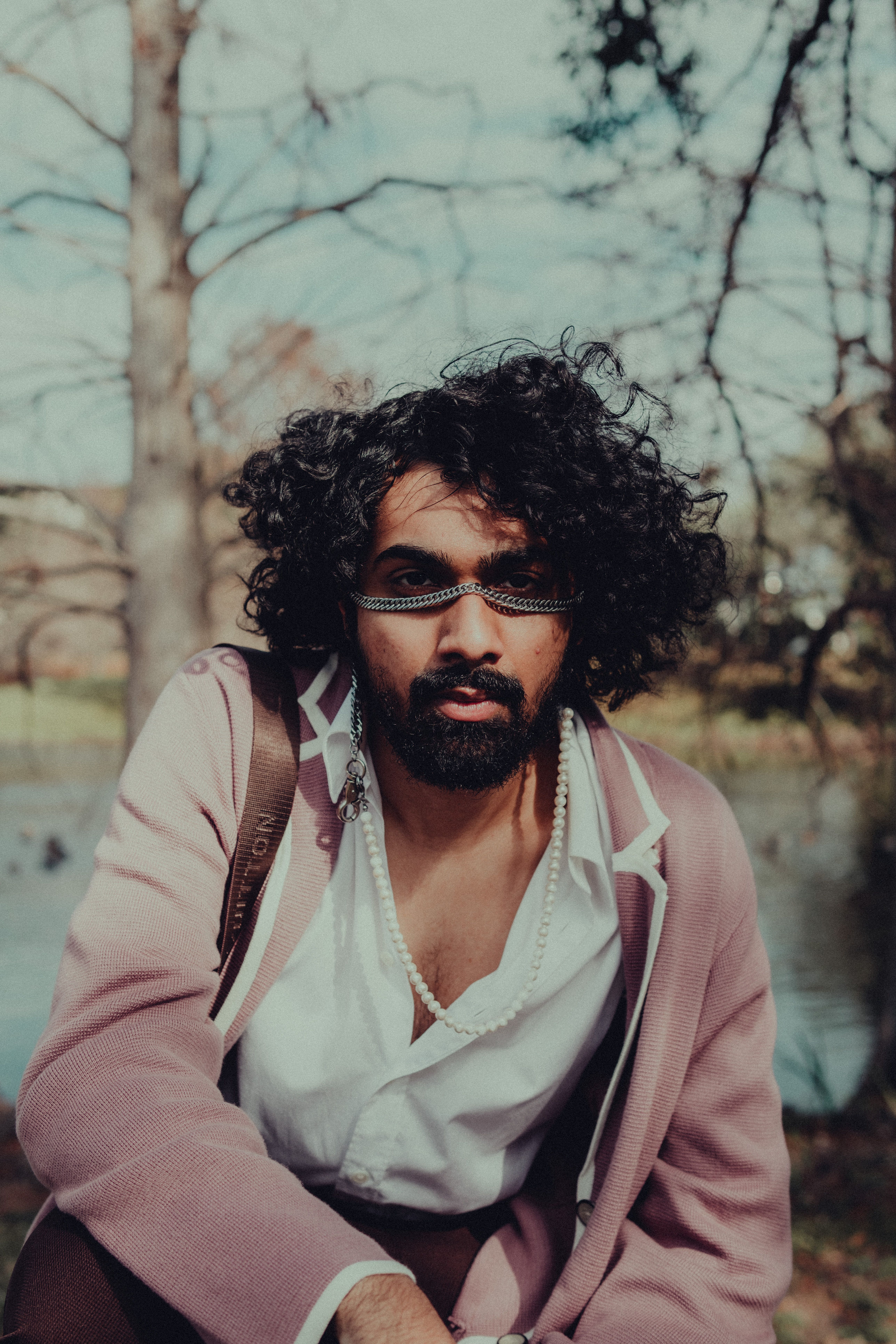 Stylish man with curly hair in a pink outfit sitting by a lake with leafless trees.