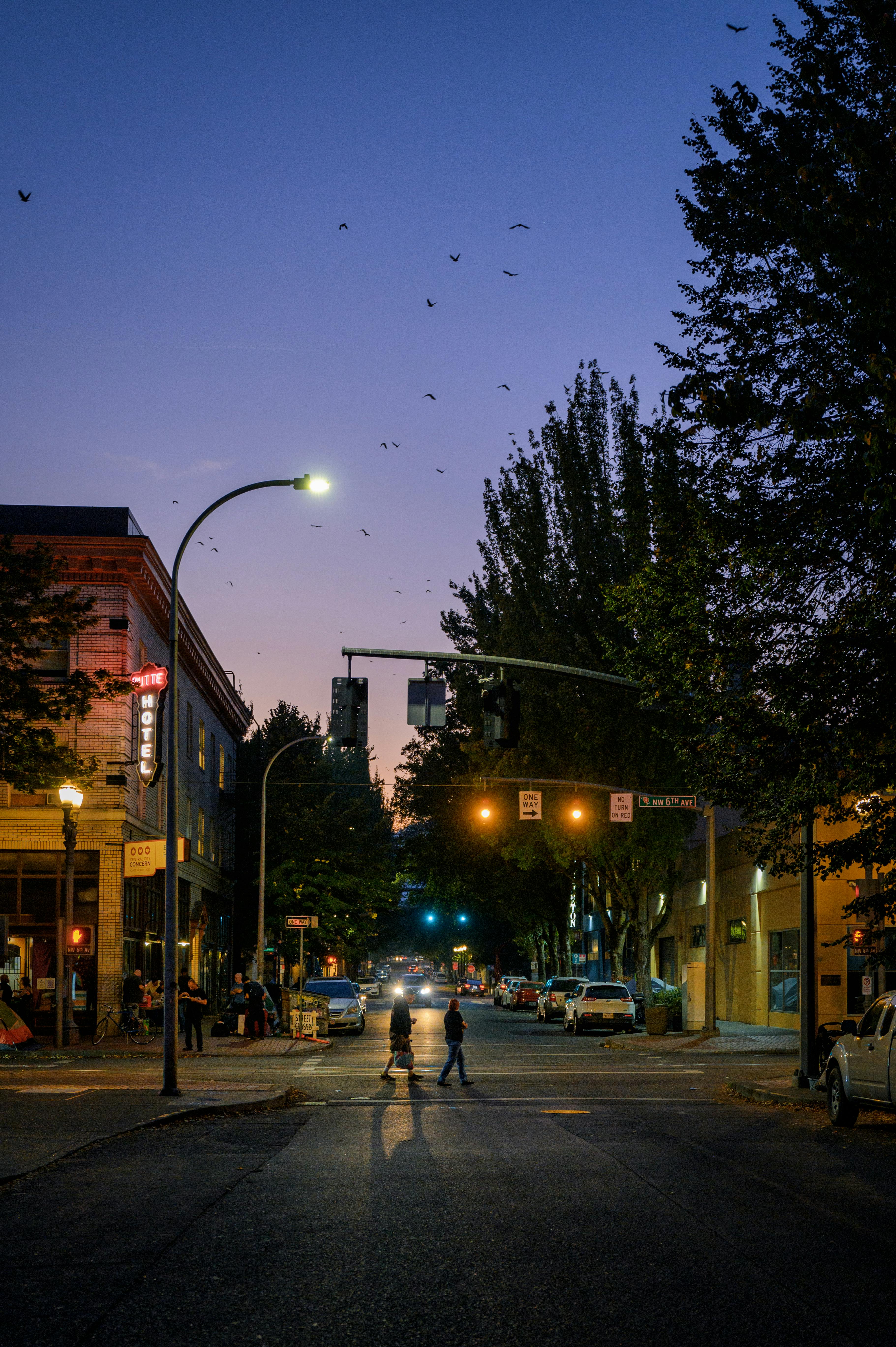 Nighttime urban street scene with silhouettes of two people crossing, lamp posts, and buildings.