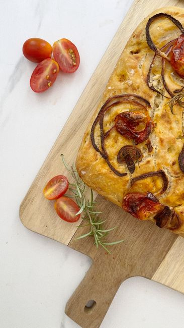 A golden, dimpled sourdough focaccia in a baking pan, glistening with olive oil and scattered with flaky salt and fresh rosemary sprigs, cut into squares.