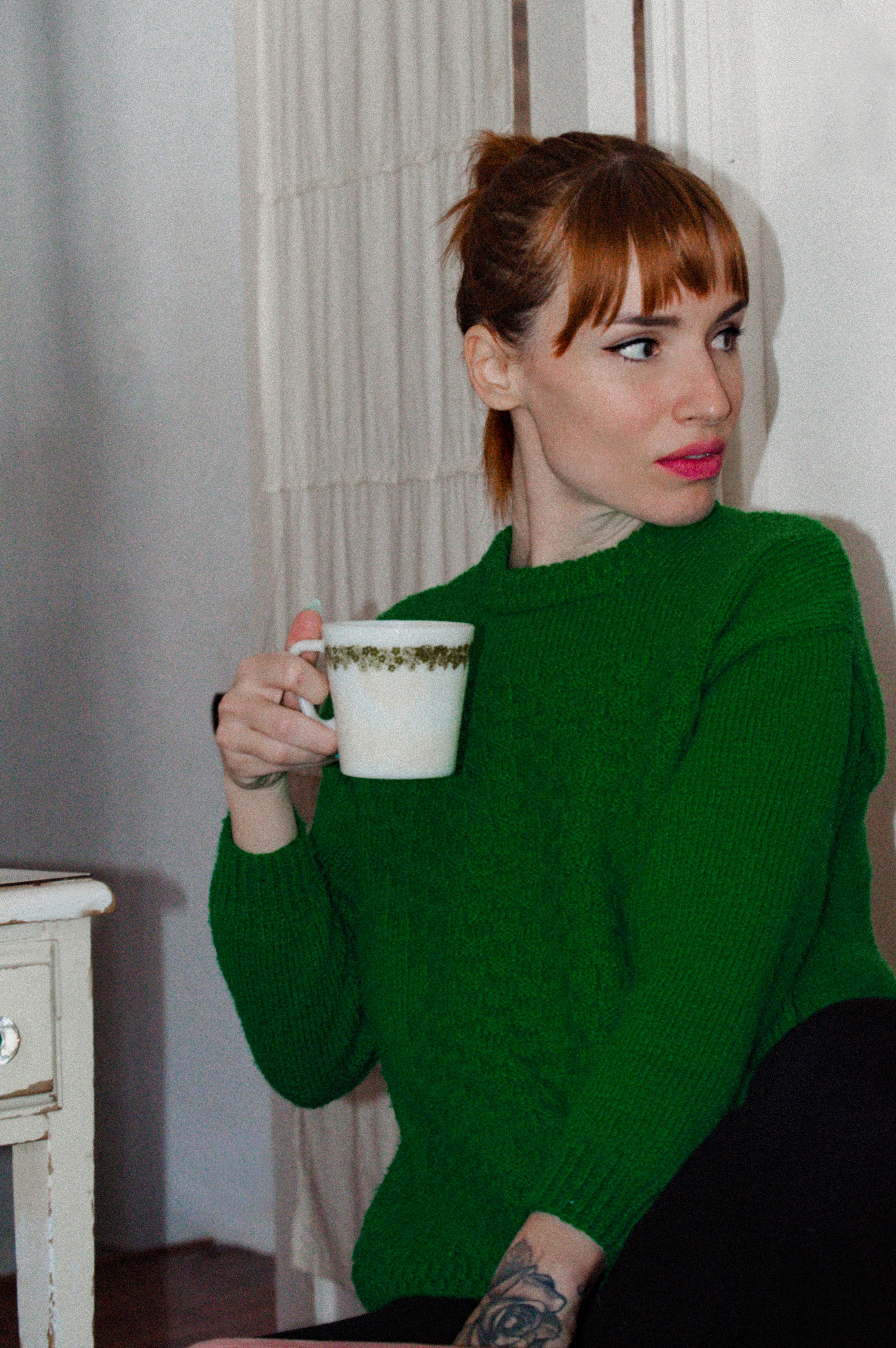 A woman with red hair in a green sweater enjoys coffee at home, creating a cozy atmosphere.