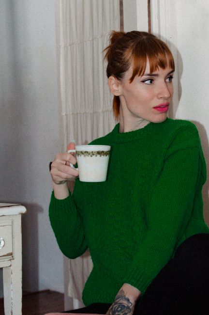 A woman with red hair in a green sweater enjoys coffee at home, creating a cozy atmosphere.