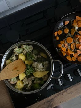 Top view of a stove with vegetables like courgettes, cauliflower, and carrots cooking in pots and pans.