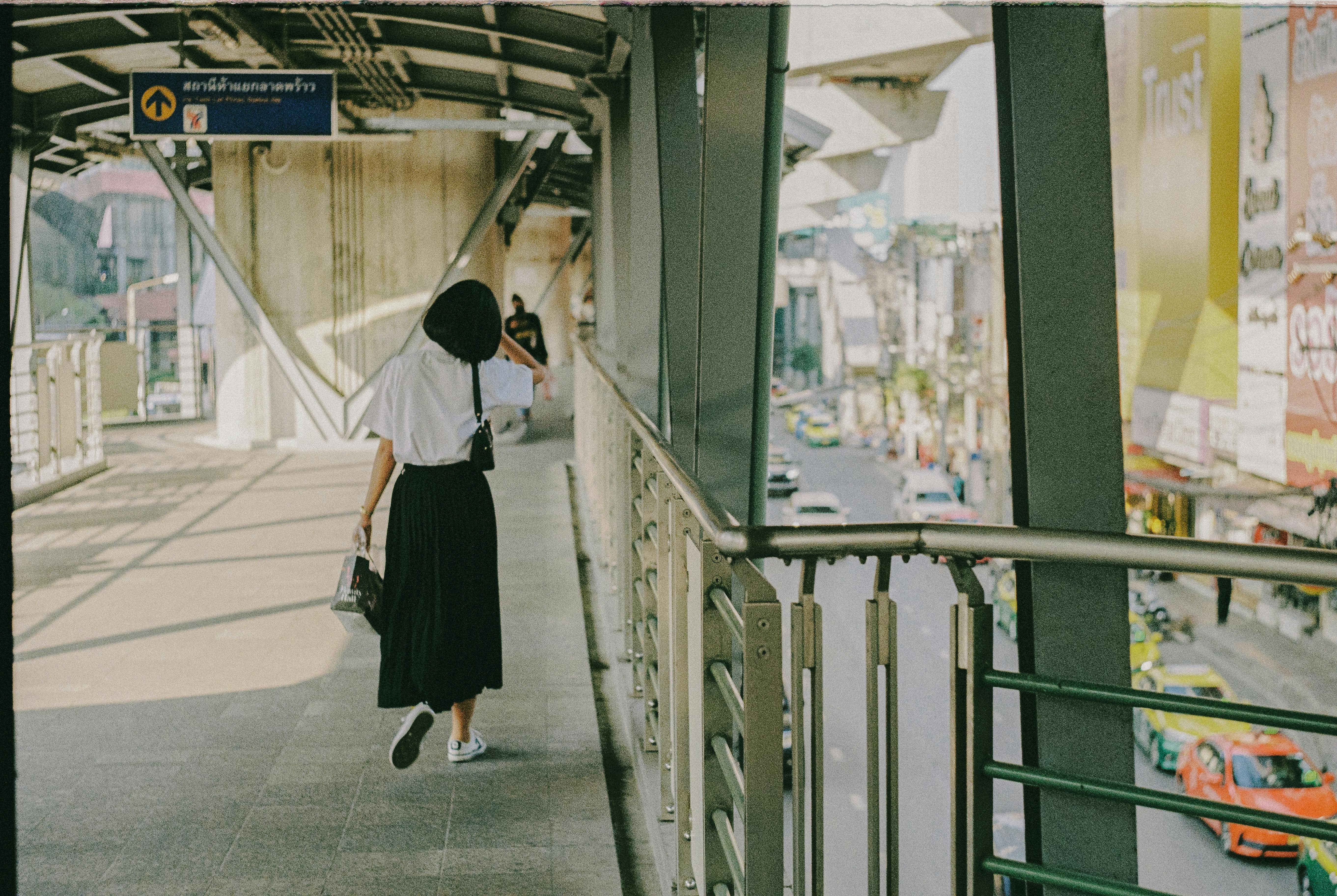 A woman walks on a city bridge pathway in Bangkok, Thailand, with urban scenery visible.