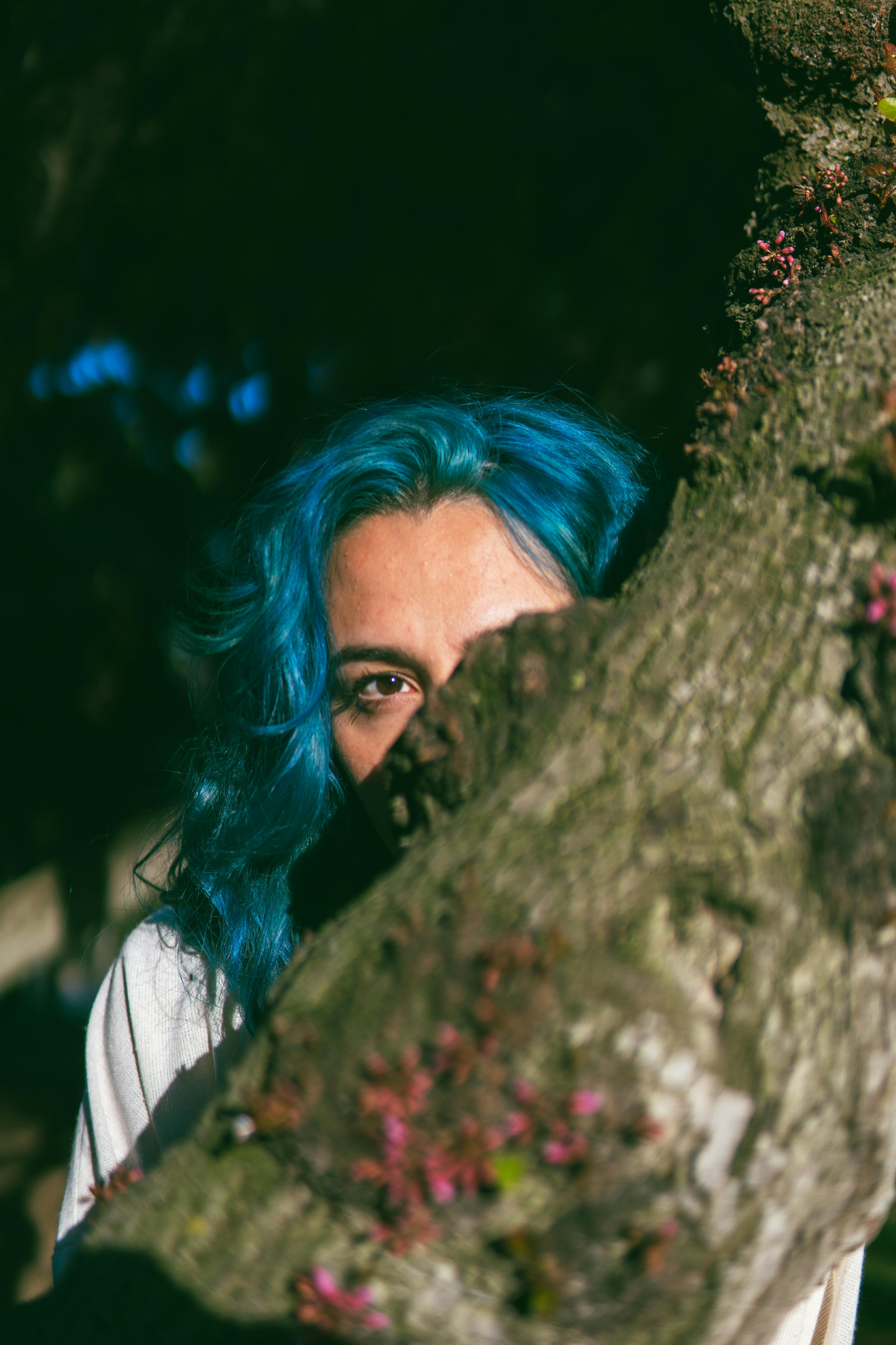 A vibrant portrait of a woman with striking blue hair peeking from behind a tree in nature.
