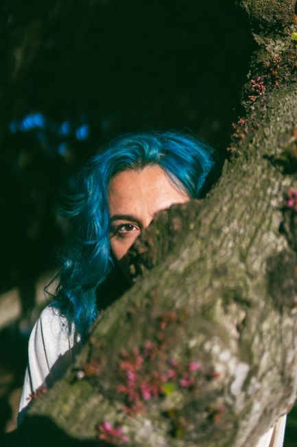 A vibrant portrait of a woman with striking blue hair peeking from behind a tree in nature.