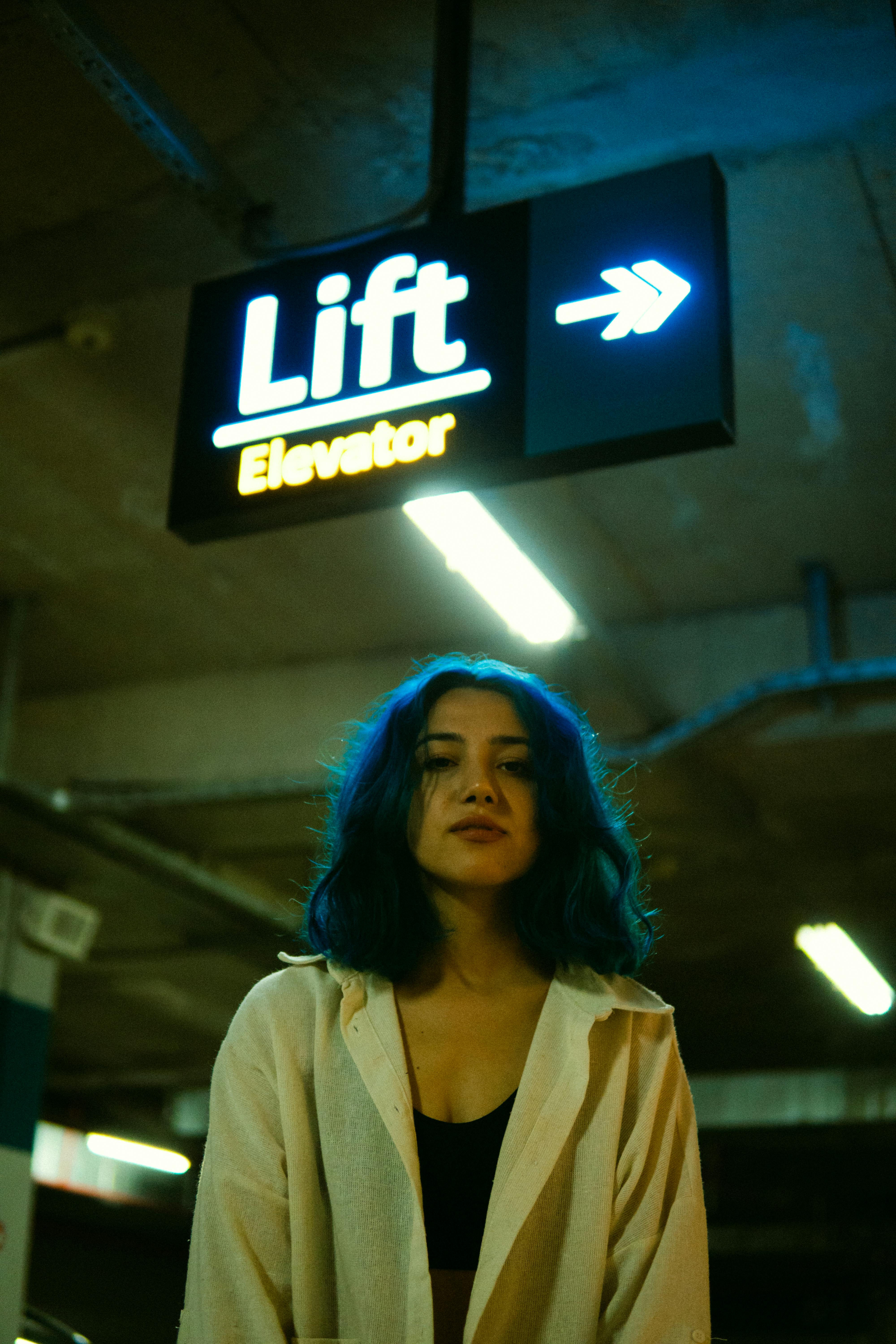 A young woman with blue hair stands under a glowing lift sign in a dimly lit parking area.