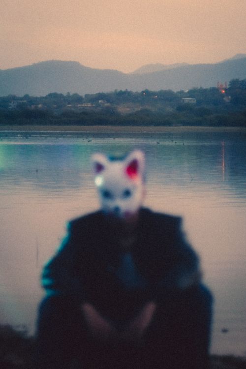 Free A person with a fox mask sits near a serene lakeside during twilight, creating a mystical atmosphere. Stock Photo