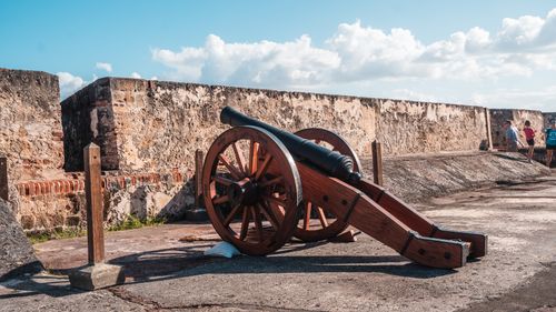 Free A historic cannon displayed at an old stone fortress with tourists passing by on a sunny day. Stock Photo