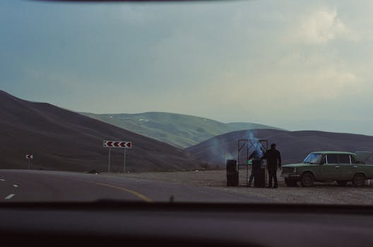 A scenic drive view with a classic car and smoky roadside BBQ under a cloudy sky.