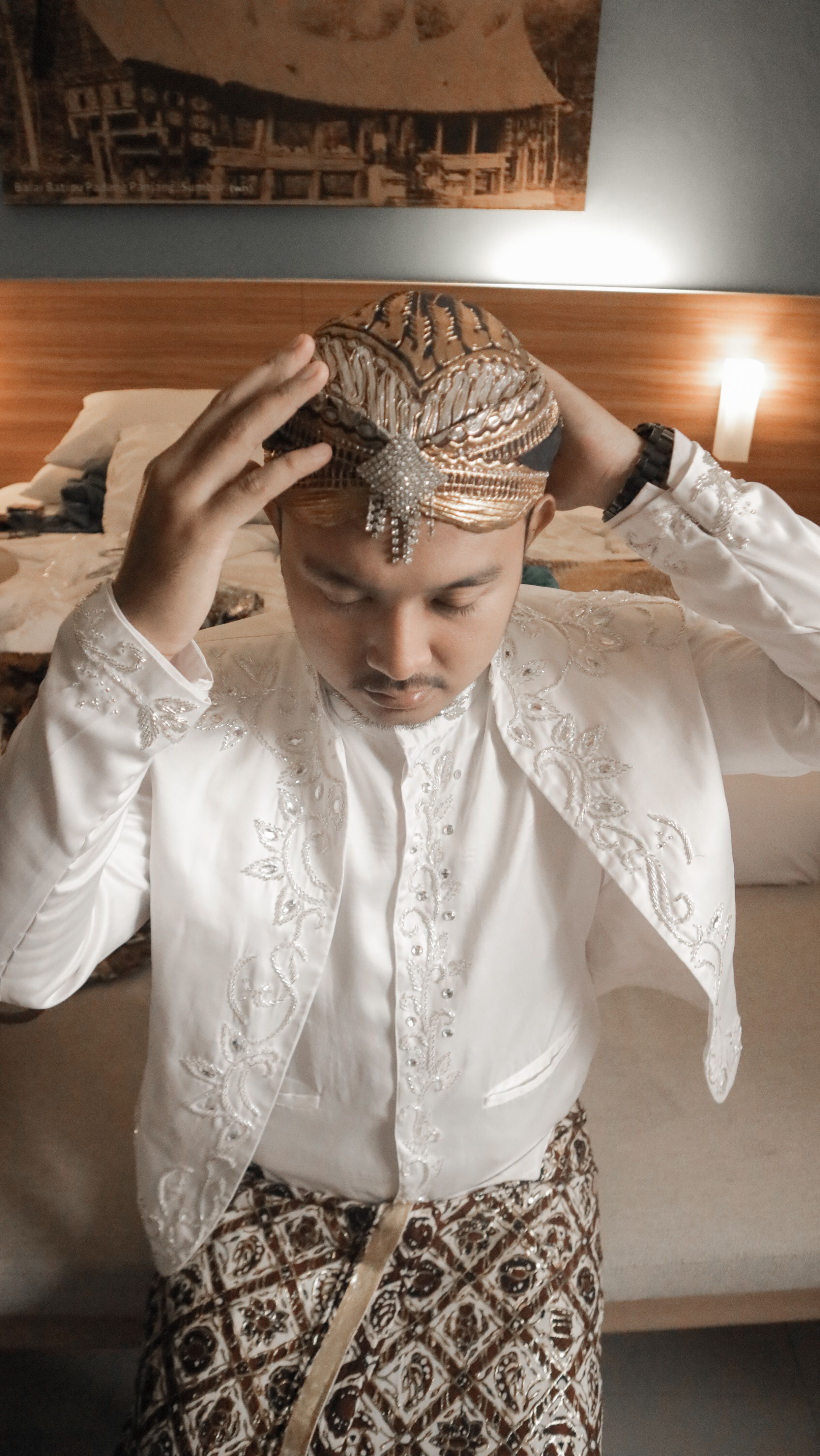 Young Indonesian groom adjusting headpiece in elegant traditional attire in a bedroom setting.